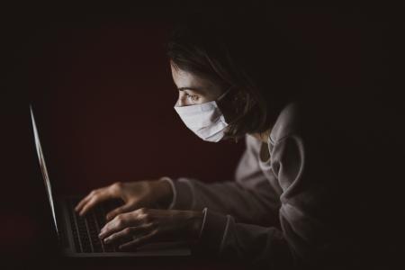 A person in a surgical mask leans over their computer in the dark.
